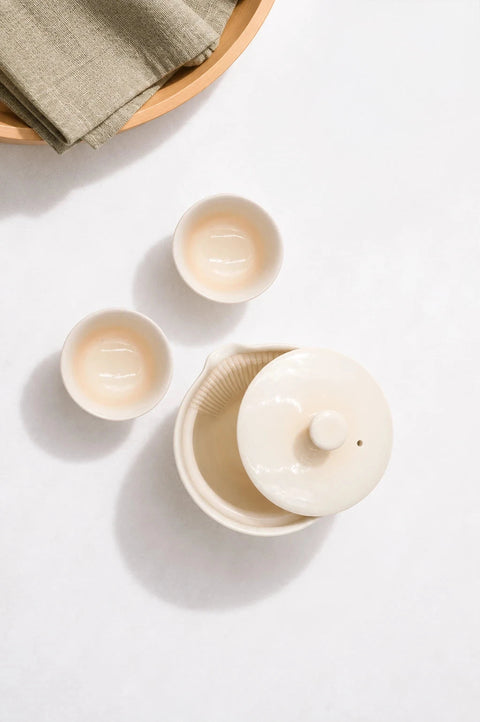 A ceramic tea set in a warm cream or off-white glaze, including a lidded teapot or tea burner, two small cups, and a folded neutral fabric beside them, arranged on a light surface with soft shadows.