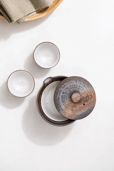 A ceramic tea set arranged on a white surface, featuring a round lidded pot with a rustic brown and gray glaze, two small white cups with thin brown rims, and a folded beige cloth at the left edge.