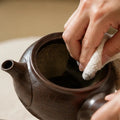 A close-up of a person's hands using a white microfiber cloth to clean the inside of a dark brown speckled ceramic teapot.