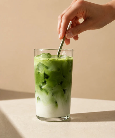 A hand holding a metal straw above a tall clear glass of layered iced matcha green tea and white milk, set on a beige surface against a warm beige background.