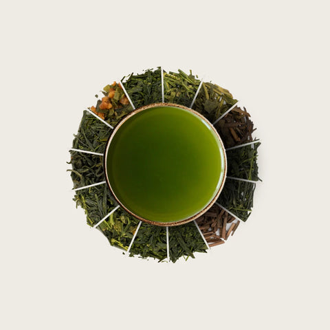 A top-down view of a ceramic cup filled with bright green tea, surrounded by a ring of loose-leaf tea blends in various shades of green and brown, set against a solid off-white background.